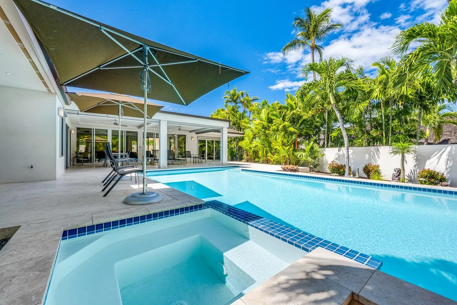 A luxury Maui pool and spa with travertine deck, umbrellas and palms under a clear blue sky